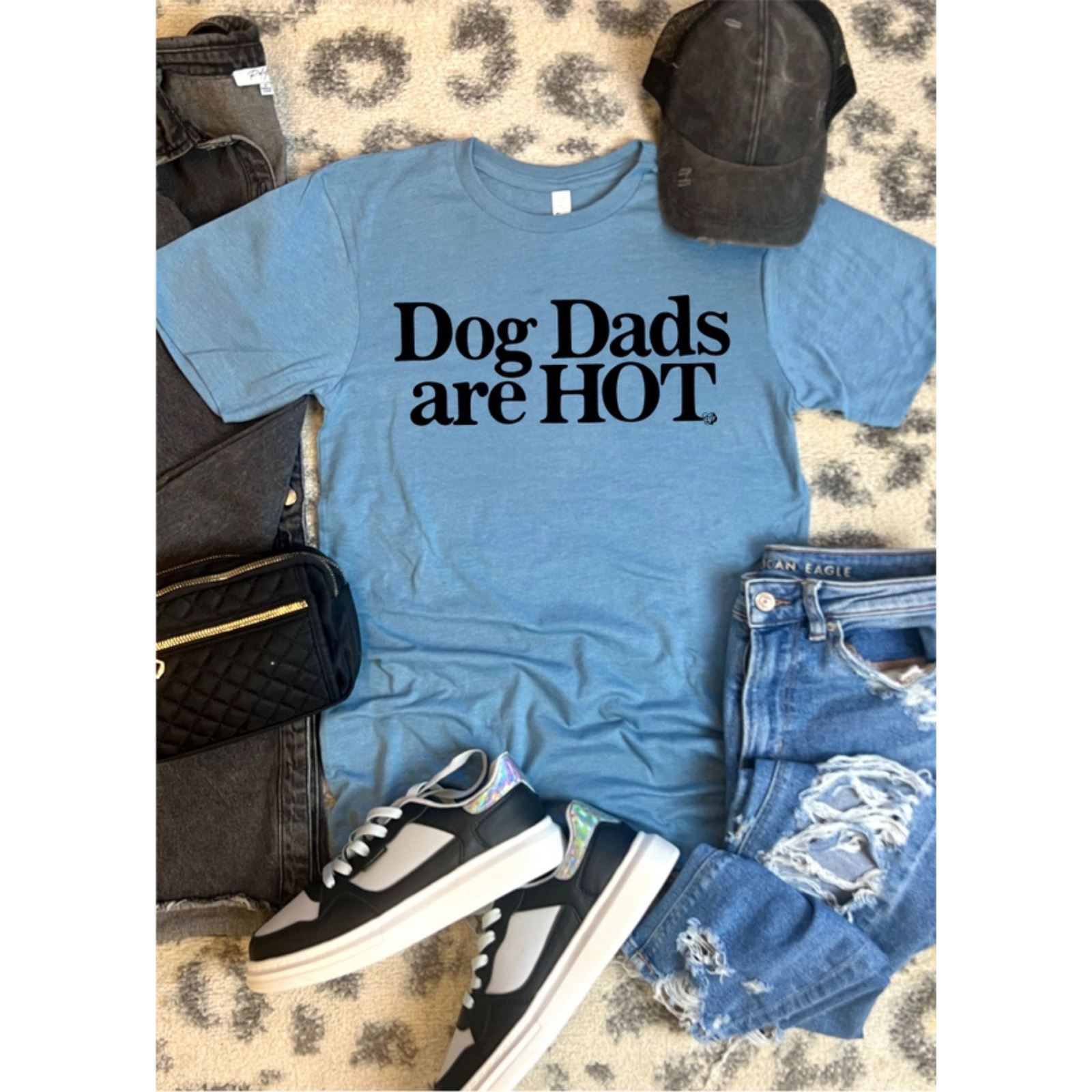 A styled flat-lay featuring a light blue graphic t-shirt with the text 'Dog Dads are HOT' in black. The t-shirt is surrounded by a matching outfit including distressed denim shorts, black sneakers, a black cap, and a black quilted bag, all arranged on a leopard-print rug.