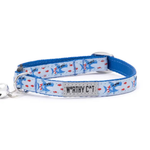 A blue fabric pet collar with a light blue patterned section featuring cartoon cats and red hearts, a silver metal buckle, and a silver bell on the left. A central label reads 'WORTHY CAT'.