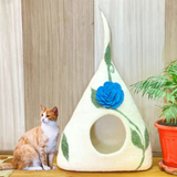 A tall cone-shaped cream felt pet cave with a large blue flower and green vines design. A white and orange cat sits next to the bed.