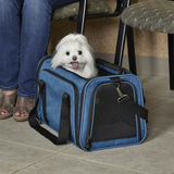 A small white dog sits in a blue pet carrier bag with black mesh and straps, placed on a tiled floor next to a person wearing jeans and sandals, with chairs visible in the background.