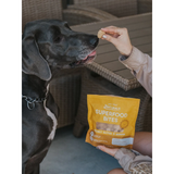 A person holds a yellow bag of Naturals Superfood Bites dog treats while offering a treat to a large black dog in a wicker patio setting.