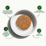 A round brown pet food disc sits in a white bowl on a light gray background, surrounded by green circular icons and text highlighting product benefits: humanely raised meat and organs, complete and balanced recipe, limited digestible ingredients, and grain and filler-free formula.