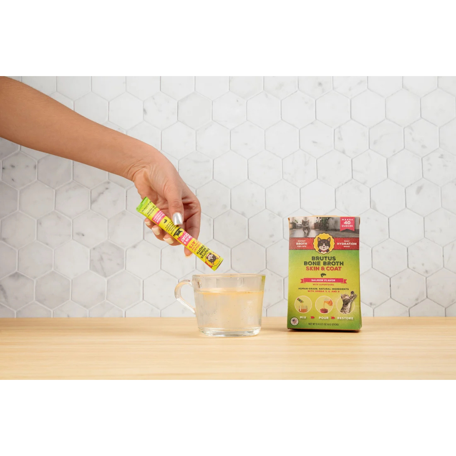 A hand holding sticks over a glass of water, with the product packaging box standing on a wooden table against a white hexagonal tile wall.