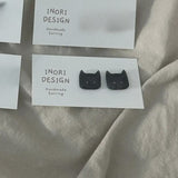 Black cat-shaped earrings on a 'INORI DESIGN' card with a beige fabric background