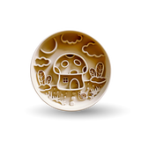 A round ceramic plate with a whimsical embossed design of a mushroom house, clouds, and plants in warm brown and cream tones, shown on a white background with a soft shadow.