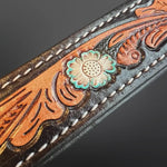 Close-up of a tooled leather strap with floral and scroll embossing, white stitching, and a turquoise-and-cream flower concho on a dark gray background.