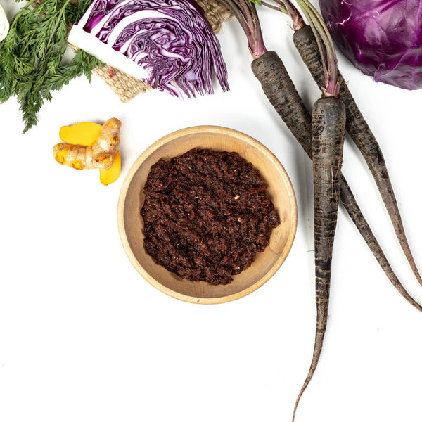 Wooden bowl with dark paste, surrounded by purple cabbage, ginger, and other vegetables on a white background