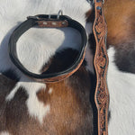 A black leather dog collar and tooled leather wavy collar laid on a brown and white cowhide background. The collar has a black interior, silver metal hardware, and a tooled outer edge.