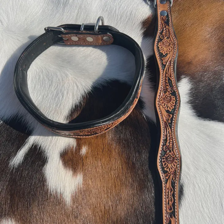 A black leather dog collar and tooled leather wavy collar laid on a brown and white cowhide background. The collar has a black interior, silver metal hardware, and a tooled outer edge.