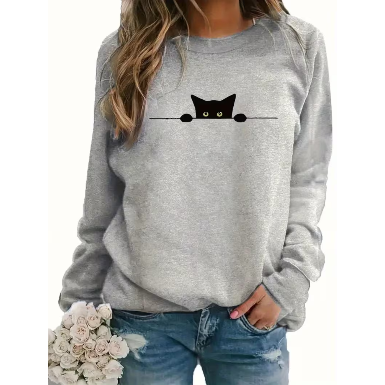 A woman wearing a light gray crewneck sweatshirt with a black cat peeking out from behind a horizontal line across the chest, paired with blue jeans and holding a bouquet of white roses.