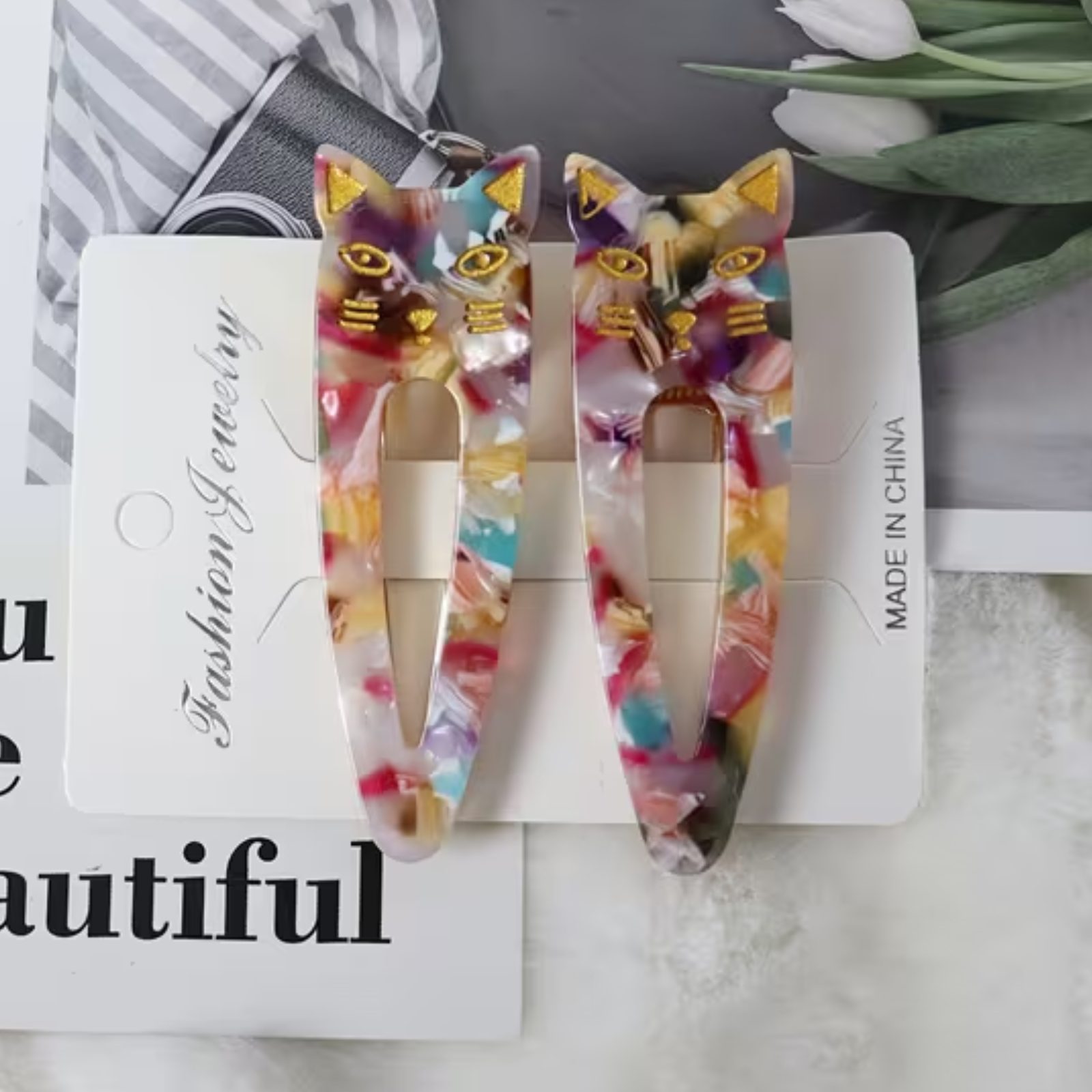 A pair of acrylic cat-ear hair clips with a colorful marbled pattern and gold cat face details, displayed on a white card