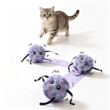 Three purple fuzzy cat toy spiders with black legs are arranged on a light purple arrow-shaped strap on a white background, with a gray tabby cat walking above them.