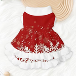 A red and white pet dress with a festive winter theme, featuring a red bodice with white snowflakes and a white ruffled hem, displayed on a white textured background with a wooden hat prop.
