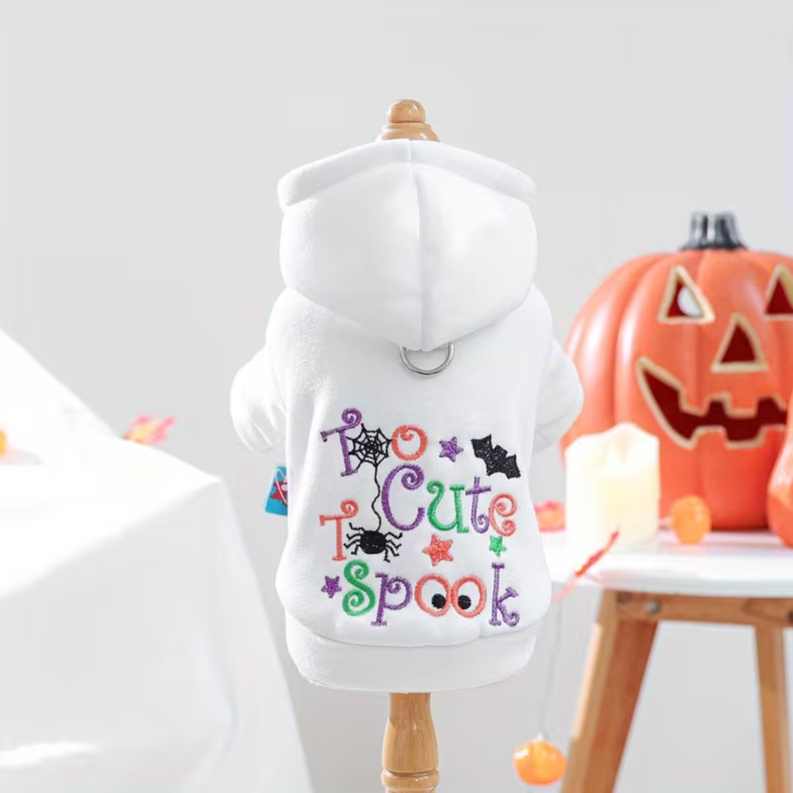 White dog hoodie with colorful 'Too Cute To Spook' text and Halloween graphics on a mannequin, with pumpkins and candles in the background.