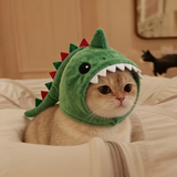 A cream-colored cat wearing a plush green dinosaur costume hood with red spikes and white teeth, sitting on a bed with white bedding.