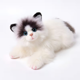 A fluffy faux-fur toy kitten lying down on a clean white background. The toy features a white body with black and gray patches on its face, ears, and tail, large blue eyes, and pink-tipped paws.