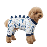 A small tan dog wearing a full-body dinosaur-print pet jumpsuit with dark blue trim and spikes, photographed on a plain white background.