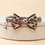 A pet collar with a decorative bow, featuring a cream-colored fabric printed with orange, brown, and black floral motifs, complete with a silver metal bell and buckle.