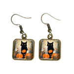 A pair of dangling earrings with square glass charms showing a black cat sitting on a pumpkin in an autumn scene, with bronze-tone fishhook wire.