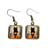 A pair of dangling earrings with square glass charms showing a black cat sitting on a pumpkin in an autumn scene, with bronze-tone fishhook wire.