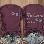 A flat-lay presentation of a burgundy t-shirt showing both front and back designs. The front features two paw prints in pastel pink and blue, while the back displays the text 'EASILY DISTRACTED BY DOGS.' and has two pastel paw prints.