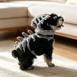 A small black and white dog wearing a gray plush dinosaur costume with a bone-look back and neck cover, standing on a light rug in a living room with a sofa in the background.