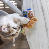 A fluffy white, gray, and brown kitten plays with a plush toy shaped like a gingerbread house and a small blue bag labeled 'Pine Snacks' on a light wood surface.