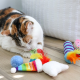 A calico cat lies on a light wood floor next to a colorful cat toy, with other toys visible in the background near a white textured cushion.