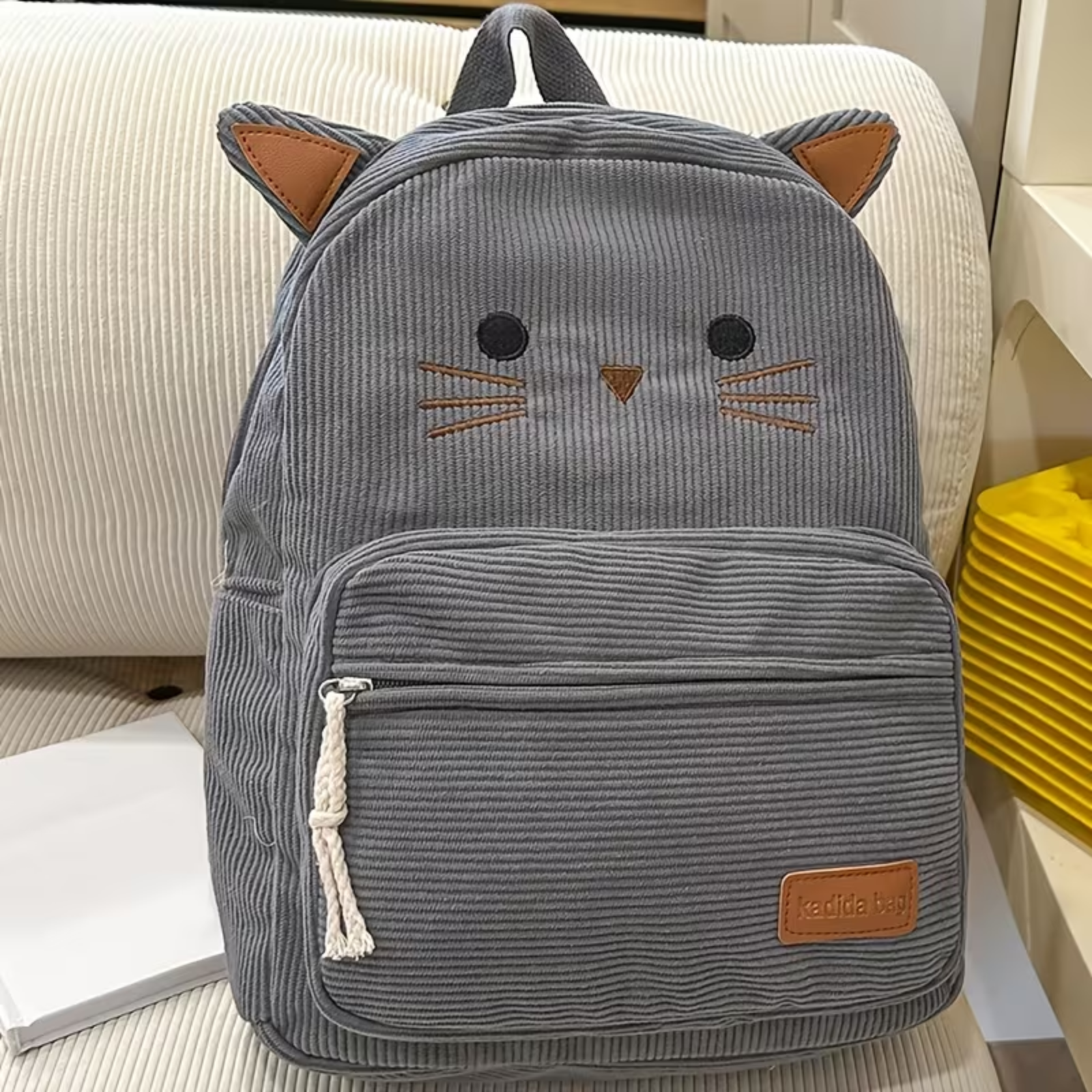 A gray corduroy cat-face backpack with brown ear patches and a small brown brand patch on the front pocket, photographed on a cream upholstered chair.