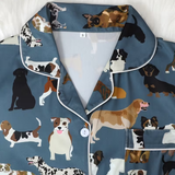 A close-up of a pajama top with a slate blue base and an all-over print of various dog illustrations. The pajama features white piping along the collar, placket, and pocket edges, and a single white button at the center front.