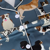 Close-up of a pajama-style top with a blue-gray satin-like fabric and an all-over print of different dog illustrations. The garment has white piping along the collar, placket, and pocket edges, plus white buttons.
