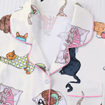 A close-up of a pajama top featuring a playful print of various cats in different poses, with pink piping along the collar and pocket edges.