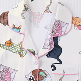A close-up of a pajama top featuring a playful print of various cats in different poses, with pink piping along the collar and pocket edges.