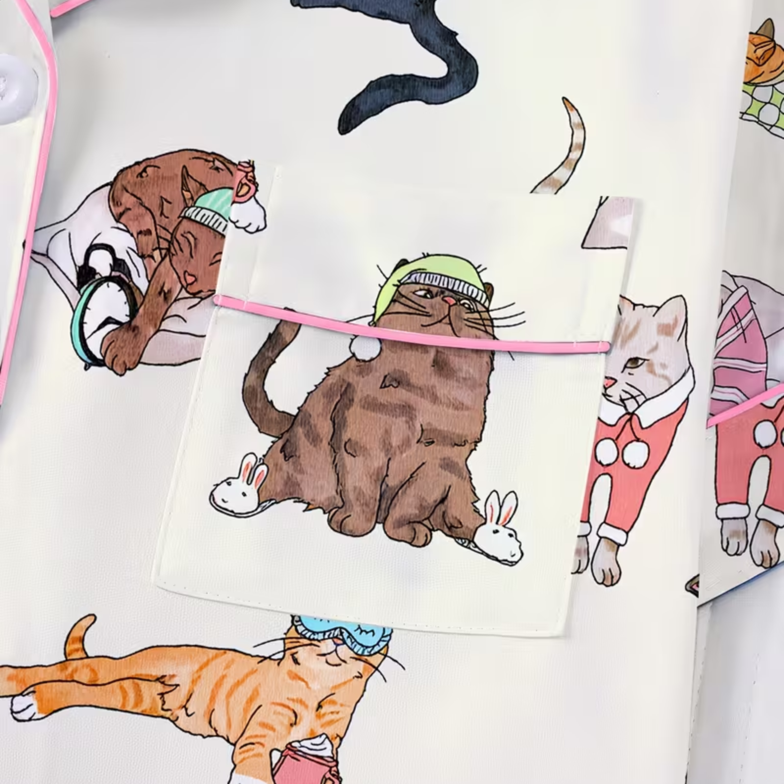 A close-up of a white fabric pajamas, printed with cute cartoon cats in different poses and outfits. A pink piping is stretched horizontally across the center of the pocket.