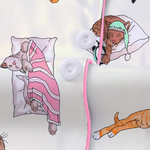 Close-up of a white button-front garment with pink trim and printed illustrations of sleeping cats in pajamas, including a pink-striped blanket and a green nightcap.