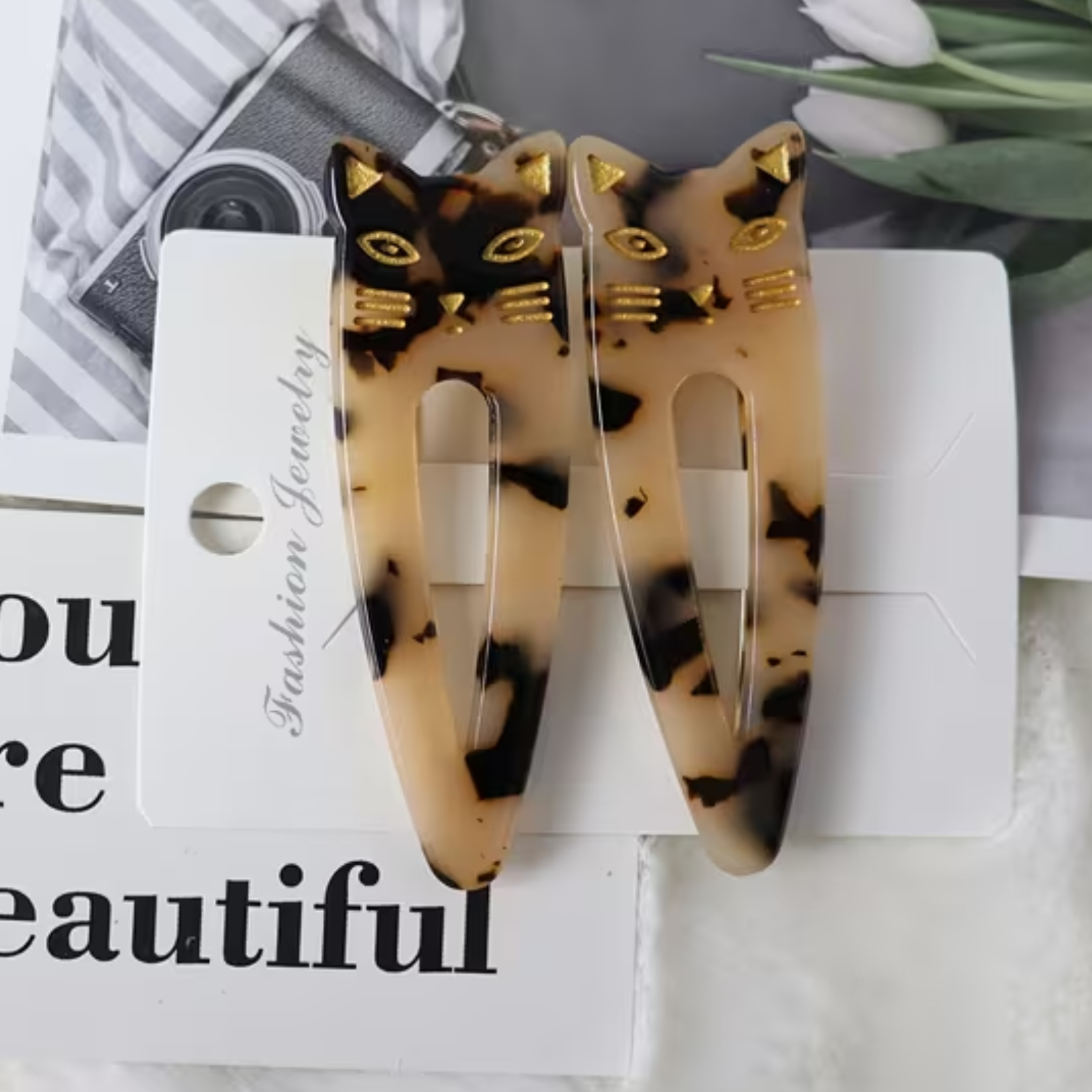 A pair of tortoiseshell cat-ear hair clips with gold eyes, whiskers, and ears, attached to a white card