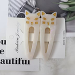 A pair of opal-white cat-shaped hair clips with gold accents, displayed on a white card