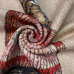 A close-up of a patterned fabric draped in a spiral, featuring a mix of red, pink, black, and beige branches with a textured beige knit fabric background.