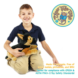 A smiling young boy in a navy polo shirt and khaki pants sits on a white background holding a plush dog toy. A circular Tiger Tales Toys logo appears in the upper right, and text at the bottom right states product safety information.