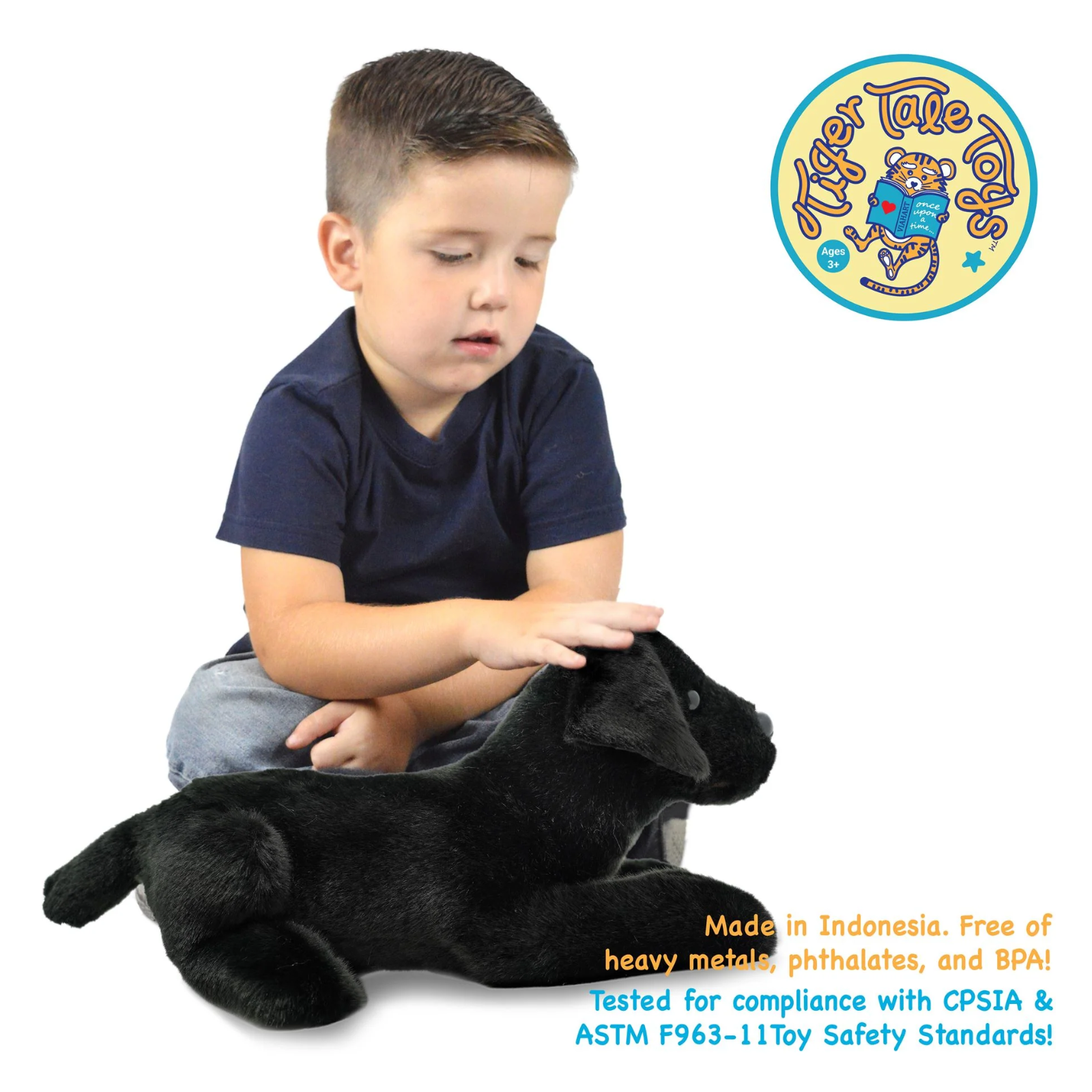 A young boy in a navy blue t-shirt and gray pants sits on a black plush stuffed animal against a white background. A circular 'Tiger Tales Toys' logo is visible in the top right, and safety text is printed at the bottom right.