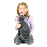A young girl with blonde hair kneeling on a white background, smiling while holding a large, realistic gray cat plush toy.