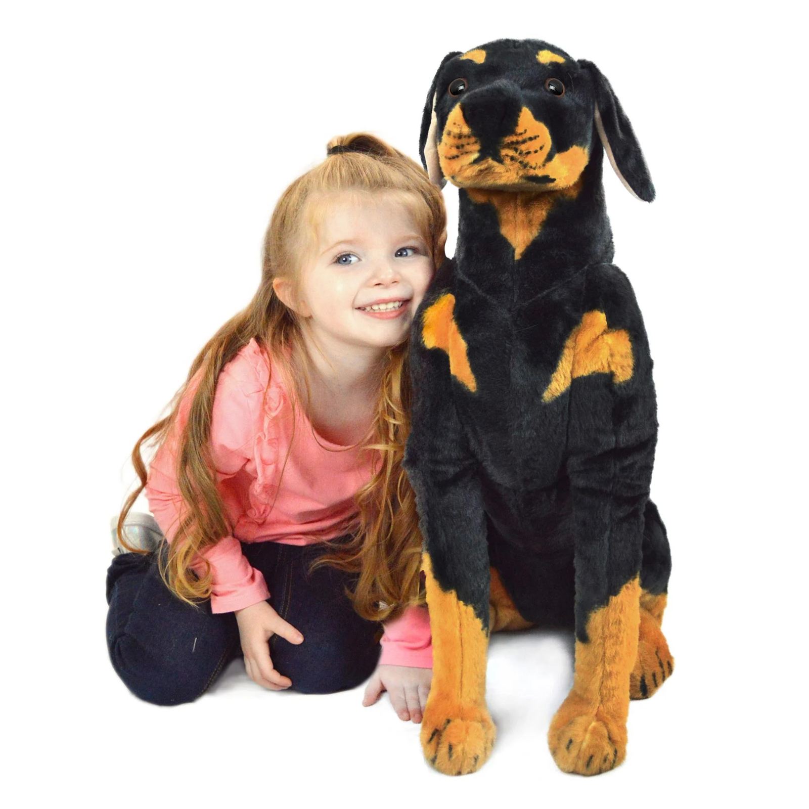 A young girl with long light brown hair sits next to a large black and tan plush dog toy against a plain white background.