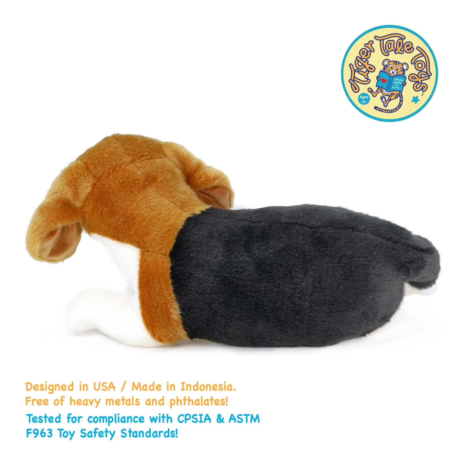A plush toy dog lying down, designed like a beagle with brown ears and head, a black body, and a white front paw. The image is on a white background with a circular 'Tiger Tale' logo in the top right and text detailing safety standards at the bottom left.