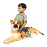 A young boy wearing a sage green polo shirt and denim shorts sits on a large, realistic plush German Shepherd dog toy against a solid white background.