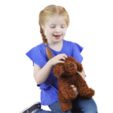 A young girl with blonde pigtails wearing a blue ruffled shirt and jeans, smiling while holding a brown curly-haired plush dog toy against a solid white background.