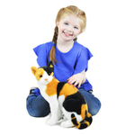 A smiling young girl with braided hair sits cross-legged on a white background, holding and petting a realistic calico cat plush toy. 
