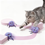 A gray tabby cat plays with three purple fluffy spider-shaped toys on a white studio background with pink geometric platforms.