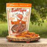 A bag of Sam's Yams Bichon Fries dog treats stands on a round wooden slice outdoors, with several orange-brown wavy fries placed in front. The background is a blurred green outdoor setting.
