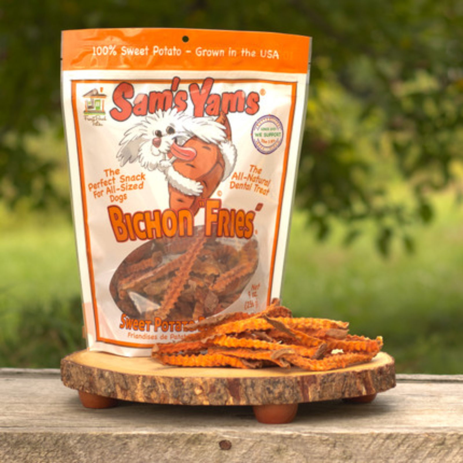 A bag of Sam's Yams Bichon Fries dog treats stands on a round wooden slice outdoors, with several orange-brown wavy fries placed in front. The background is a blurred green outdoor setting.