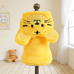 A bright yellow plush animal hood or hat displayed on a dress form in a softly lit room. The fuzzy character design has embroidered black facial features, pale yellow ear and cheek patches, and is shown against a light wall with a window, curtain, and small teddy bear in the background.